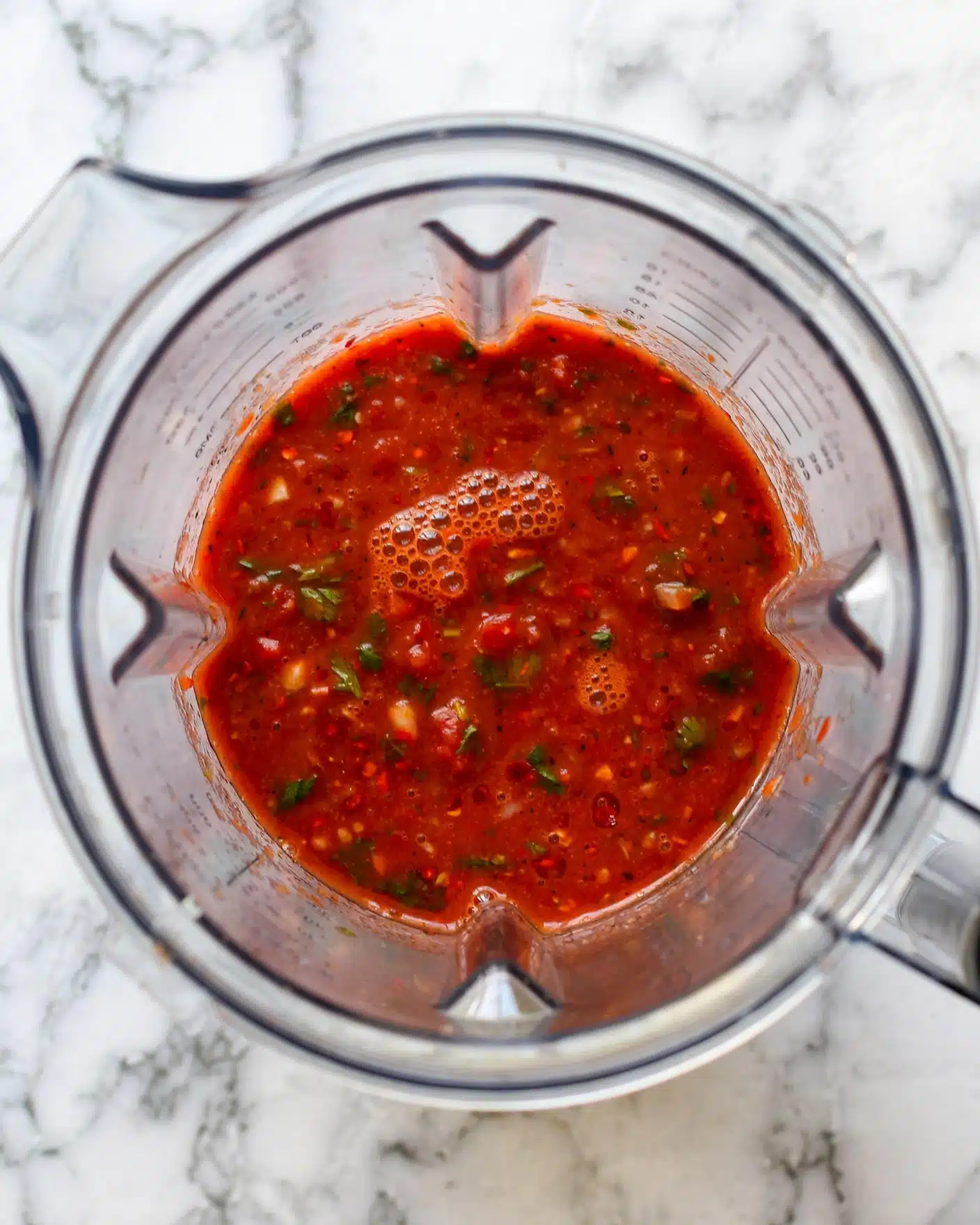 A top view of a blender container filled halfway with a smooth red sauce that has a slightly thick texture and small bubbles on the surface, showing some tiny bits of ingredients mixed inside. The clear blender sits on a white marbled surface with soft grey veins. photo taken with an iphone --ar 4:5 --v 7