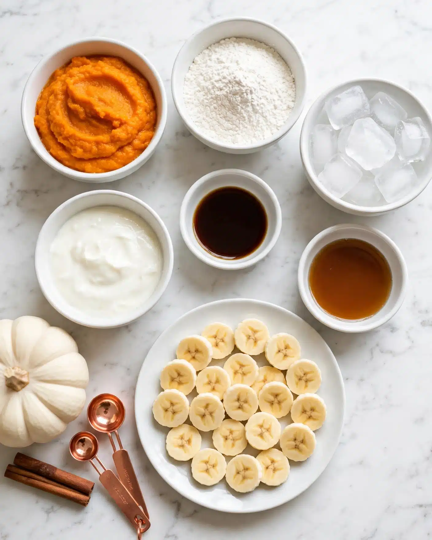 Healthy Pumpkin Spice Protein Smoothie Recipe 4 The image shows several small white bowls and a white plate arranged on a white marbled surface. There is a bowl with bright orange pumpkin puree, another with white powder, a bowl filled with clear ice cubes, and a bowl with creamy white yogurt. A small bowl contains dark brown vanilla extract, and another bowl holds a light brown syrup. The white plate holds a single layer of evenly cut round banana slices. On the side, there is a white pumpkin and two cinnamon sticks with two copper measuring spoons. The setup looks clean and organized. photo taken with an iphone --ar 4:5 --v 7