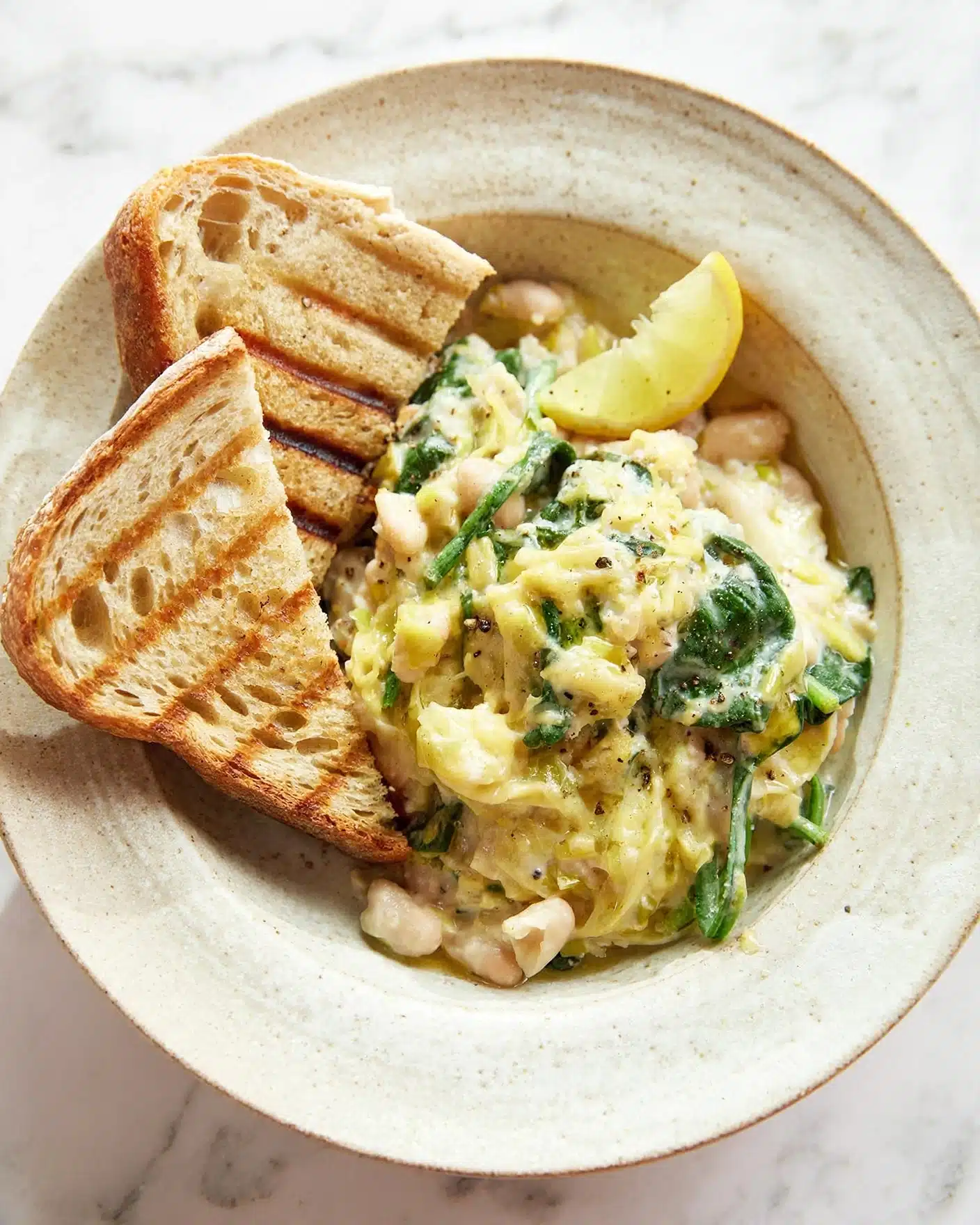 A white bowl holds a creamy mix of white beans, wilted dark green spinach, and shredded light yellow cheese, topped with a sprinkling of black pepper. On one side, there are two triangular slices of toasted bread with golden brown grill marks and a crunchy texture. A small bright yellow lemon wedge sits near the beans mixture on the edge of the bowl. The whole bowl is placed on a white marbled surface. photo taken with an iphone --ar 4:5 --v 7