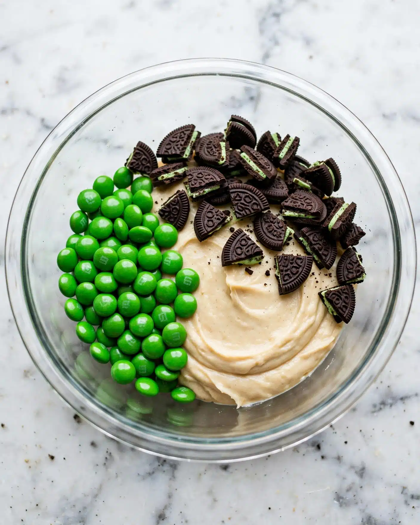 A clear glass bowl holds three main components arranged side by side. On the left, there is a pile of shiny bright green candy-coated chocolates. To the right, there is a large dollop of smooth creamy beige mixture. Covering the top right section are many small pieces of dark chocolate cookies with visible green cream filling. The bowl is placed on a white marbled surface. photo taken with an iphone --ar 4:5 --v 7