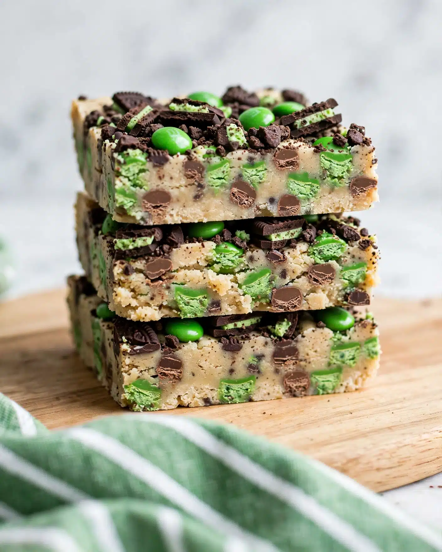 The image shows a stack of thick dessert bars with two visible layers: the base layer is light beige with a slightly crumbly texture mixed with green and brown candy pieces, while the top layer is covered with crushed dark chocolate cookie chunks and more green candy pieces embedded throughout. The bars are placed on a light wooden board, with a soft focus white marbled background, and a green cloth with white stripes is partially visible in the foreground. photo taken with an iphone --ar 4:5 --v 7
