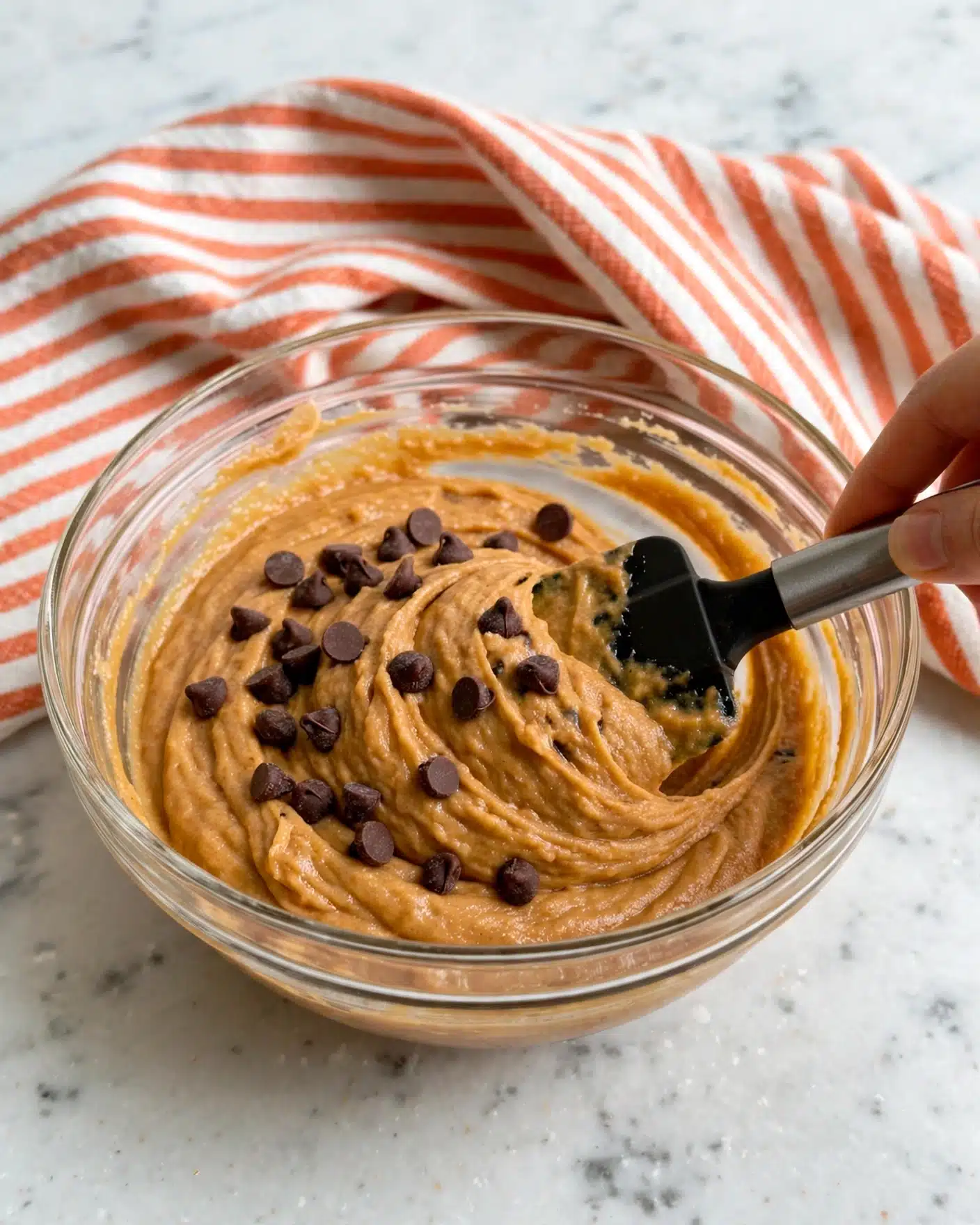 A clear glass bowl sits on a white marbled surface, filled with thick, smooth orange batter mixed with dark brown chocolate chips scattered on top. A woman's hand holds a black spatula with a silver handle, lifting and stirring the batter, creating soft swirls. The texture of the batter looks creamy and dense, with the chocolate chips sitting unevenly on the surface, adding contrast. Photo taken with an iphone --ar 4:5 --v 7