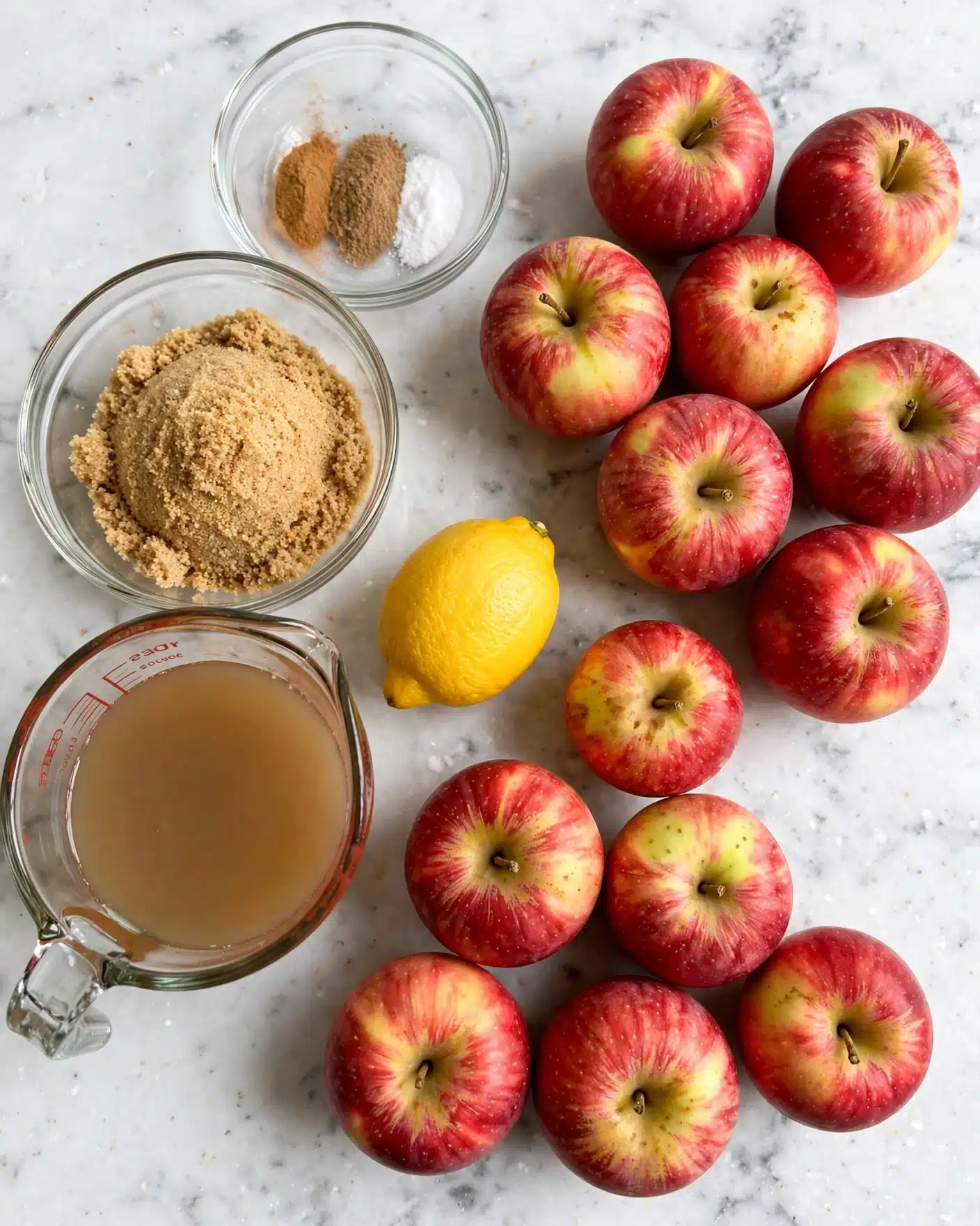 The image shows ingredients laid out on a white marbled surface, including ten red apples with yellow patches clustered on the right side. To the left, there is a clear glass measuring cup filled with light brown apple juice. Above it, a medium clear glass bowl holds light brown sugar, and to its upper left, a smaller clear glass bowl contains three spices: cinnamon, nutmeg, and baking soda, arranged side by side. A whole bright yellow lemon sits between the measuring cup and the apples. The photo taken with an iphone --ar 4:5 --v 7