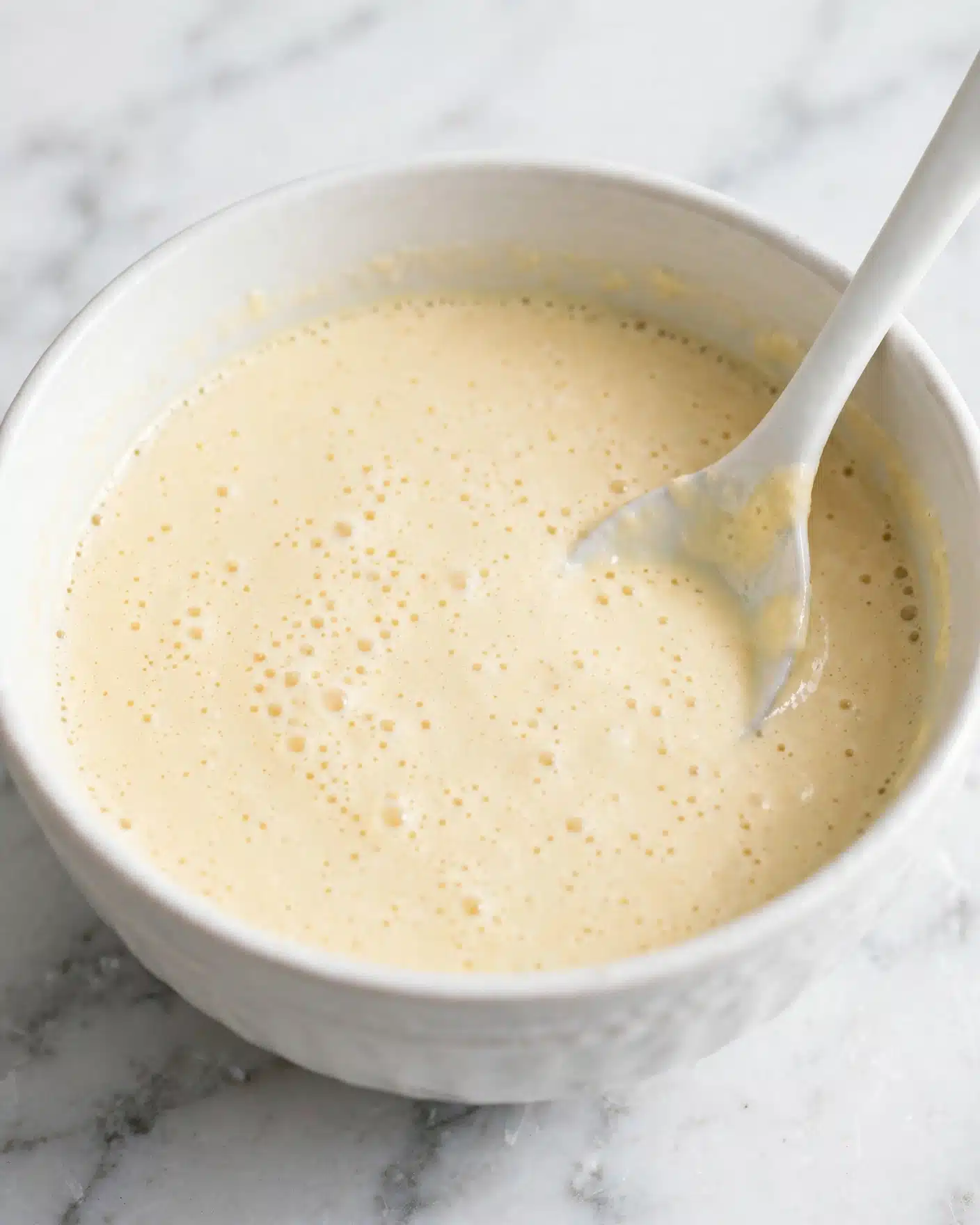 A close-up view of a white bowl with smooth, creamy pale yellow sauce inside, tiny bubbles visible on the surface to show light texture; a white spoon inside the bowl resting at an angle on the right side, partially dipped into the sauce; the bowl is placed on a white marbled texture background that contrasts softly with the sauce and bowl. photo taken with an iphone --ar 4:5 --v 7