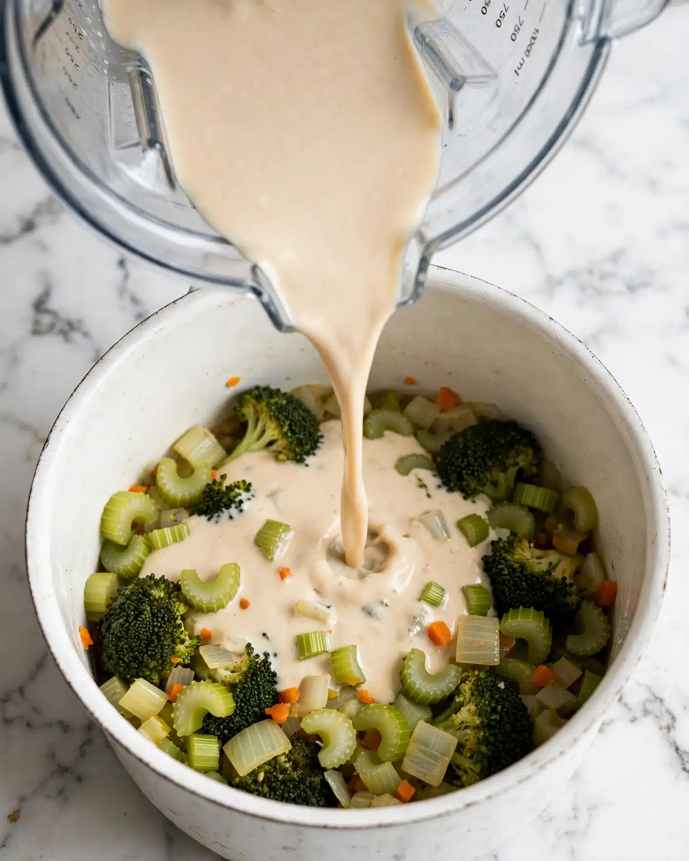 Detox Vegetable Soup with Cashew Cream and Lentils Recipe 4 A white pot filled with a mix of cooked vegetables including dark green broccoli florets, pale green celery slices, light yellow onion pieces, and small bits of carrot, all resting at the bottom. From above, a creamy light beige liquid is being poured over the vegetables from a clear blender jar, creating a smooth layer on top. The pot sits on a white marbled surface, and the scene captures a close-up view focusing on the pouring action. photo taken with an iphone --ar 4:5 --v 7