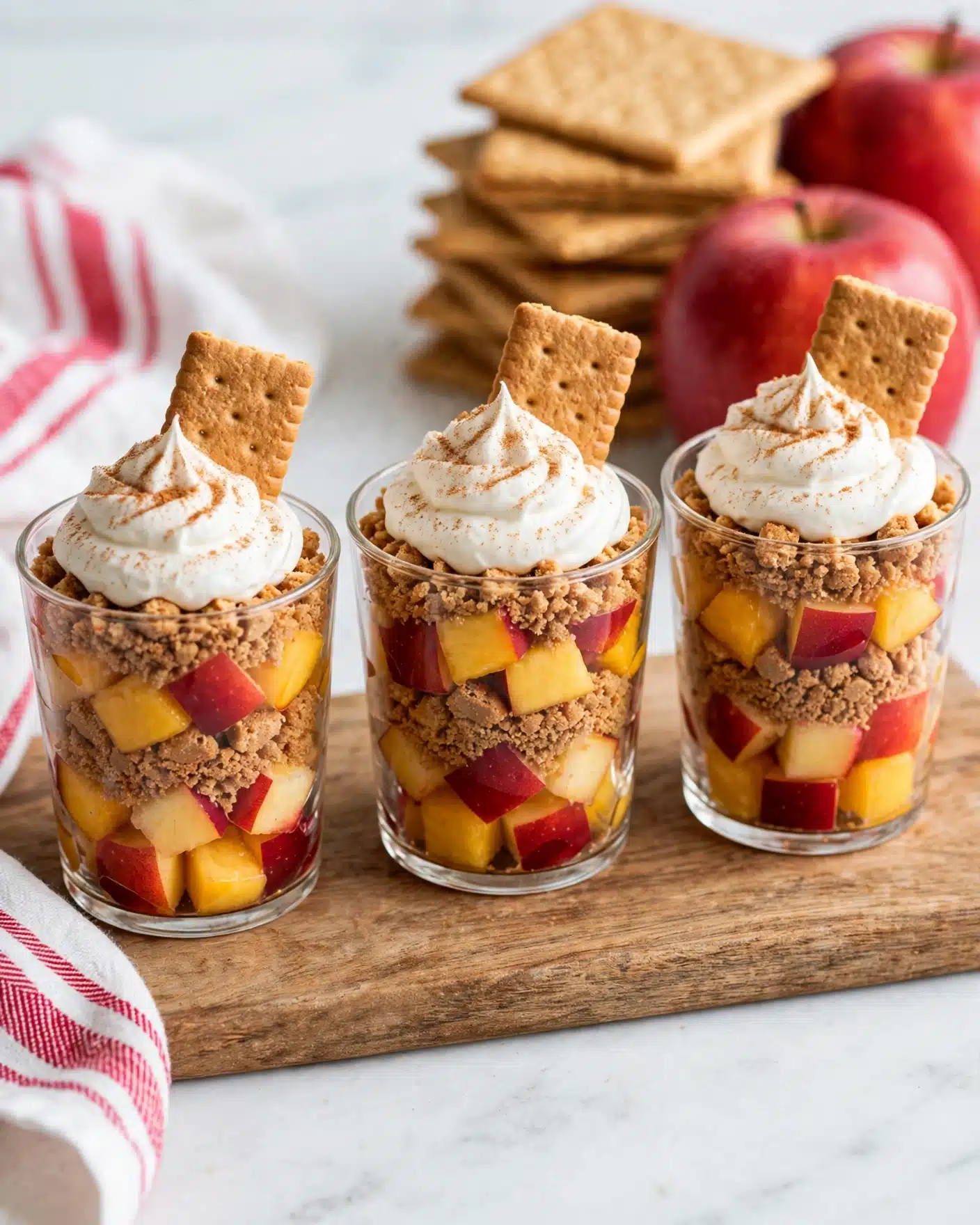 Apple Pie Shots Recipe 5 Three clear glass cups are filled with a layered dessert placed on a wooden board. Each cup has diced red and yellow peaches at the bottom, followed by a layer of brown crumbly cookie pieces, more peach chunks, and another cookie layer. They are topped with white whipped cream swirled with a sprinkle of light brown cinnamon powder, and a rectangular cookie piece sticks upright in the whipped cream. In the background, red apples and a white cloth with red stripes are visible on a white marbled surface. photo taken with an iphone --ar 4:5 --v 7