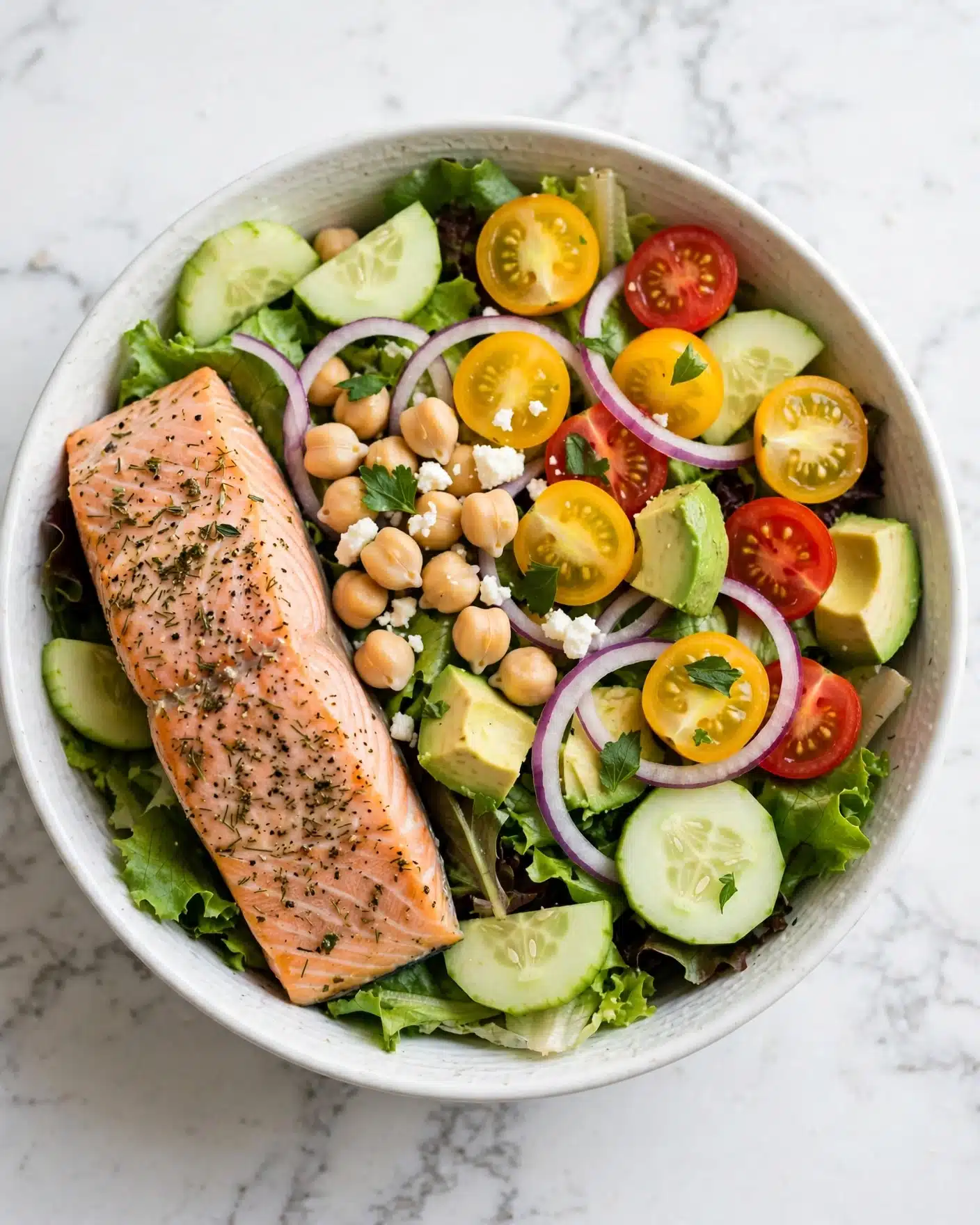 Mediterranean Salmon Quinoa Bowl with Lemon-Herb Marinade and Tzatziki Recipe 5 A white bowl filled with a colorful layered salad starts with fresh green leafy lettuce at the base. On top, there are light beige chickpeas scattered around with small bits of white crumbly cheese sprinkled over them. Bright yellow and red cherry tomato halves are spread across, along with sliced pale green cucumber and chunks of light green avocado. Thin rings of purple onion add contrast. A large piece of cooked pink salmon, seasoned with herbs and black pepper, lies on one side. Small green herb leaves are scattered on top, all placed on a white marbled surface. Photo taken with an iphone --ar 4:5 --v 7