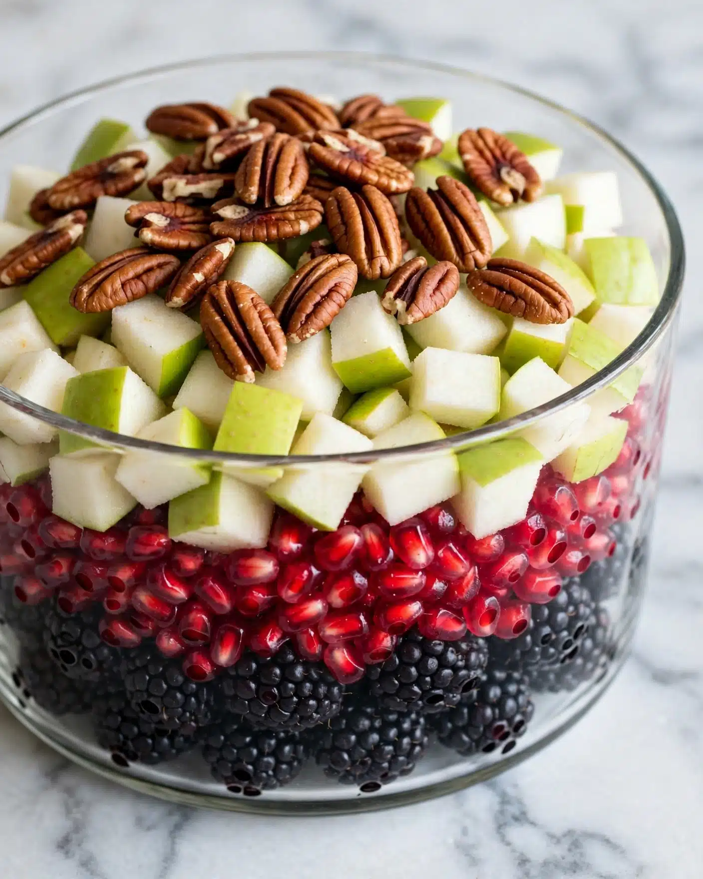 A transparent bowl filled with a layered mix of fresh fruit and nuts sits on a white marbled surface. The bottom layer consists of dark, bumpy blackberries and smaller dark berries. Above that, there is a layer of bright red pomegranate seeds, adding a shiny and juicy texture. The next layer contains small, chunky pieces of green and white fruit, likely pears or apples, showing a crisp and juicy appearance. Scattered on top are medium brown pecan nuts with visible ridges and a rough texture, providing a crunchy contrast. The mix is colorful and fresh, showing a variety of textures from smooth and juicy to rough and crisp. Photo taken with an iphone --ar 4:5 --v 7