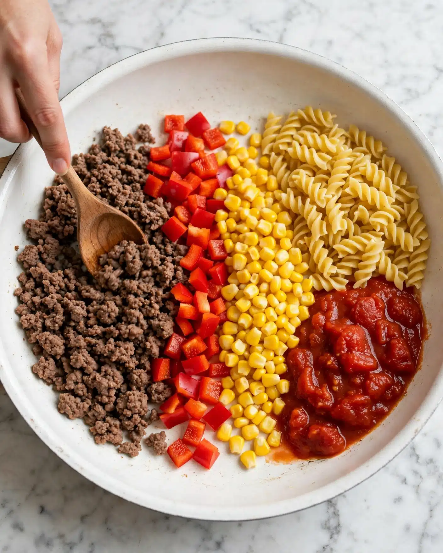 A white pan with a thick rim holds five main ingredients, each layered side by side. Starting from the left, there is browned ground meat with a crumbly texture, then fresh bright red chopped bell peppers in small cubes in the center, next to a pile of shiny cooked yellow corn kernels, a mound of yellow cooked spiral pasta filling the top right side, and finally a portion of chunky red tomato sauce with visible bits of tomato at the bottom right. A woman's hand is stirring the meat with a wooden spoon. The pan sits on a white marbled surface. photo taken with an iphone --ar 4:5 --v 7