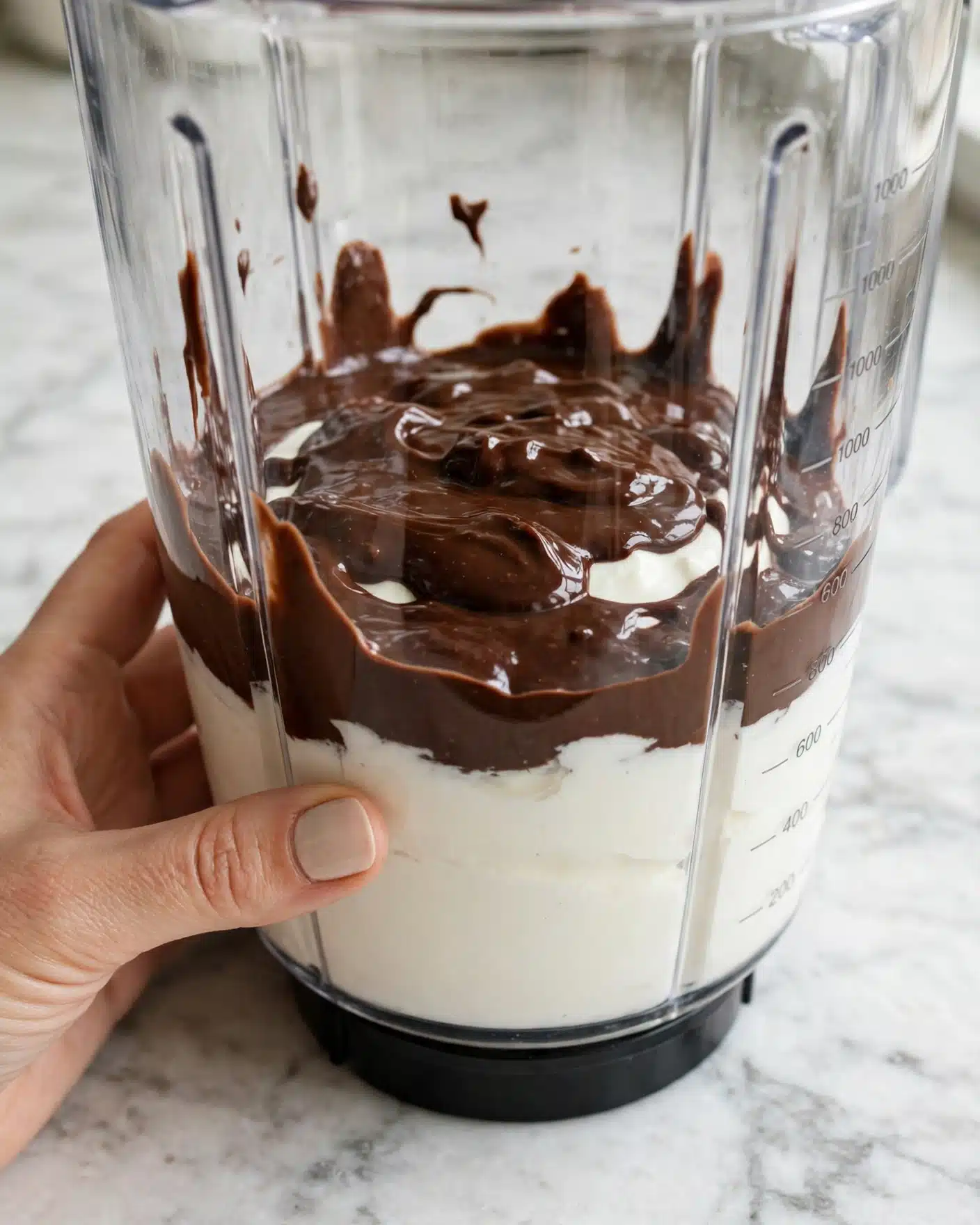 Cottage Cheese Chocolate Mousse Recipe 4 Inside a clear blender container, there is one layer of white thick yogurt at the base, topped with a smooth, shiny dark brown chocolate sauce spread unevenly on top. The container is placed on a white marbled surface, and near the blender there is a woman's hand holding it steady. photo taken with an iphone --ar 4:5 --v 7