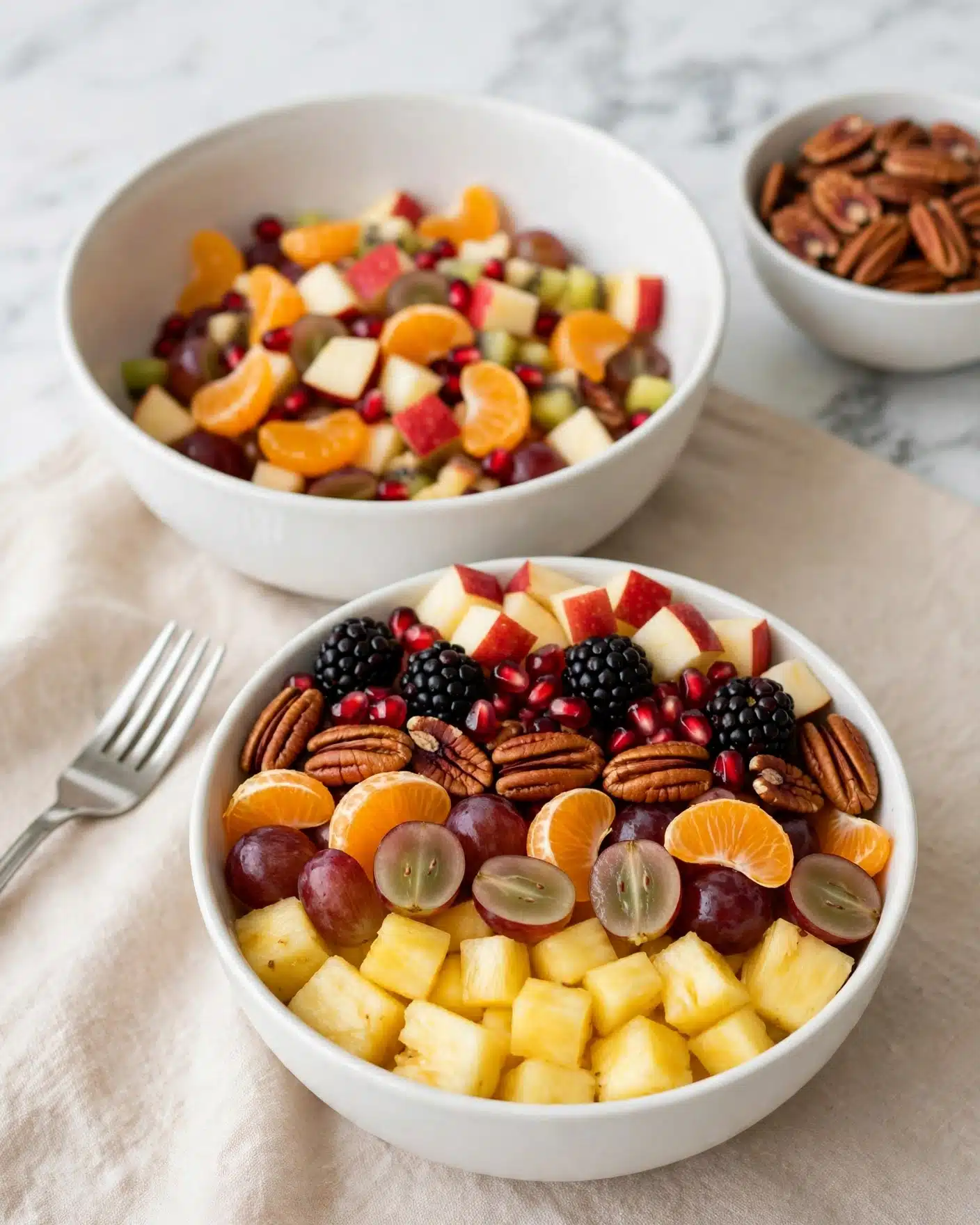 A white bowl is filled with a colorful fruit salad showing three main layers: the bottom layer has light yellow pineapple chunks, the middle layer includes red grapes cut in half and small bright orange mandarin pieces, while the top layer mixes shiny pecan nuts, large blackberries, red pomegranate seeds, and apple cubes with red skin. The bowl is placed on a soft beige surface with a silver fork to the left. A larger white bowl filled with chopped fruits is behind it, and another white bowl with pecans is seen to the top right. The background is a white marbled texture photo taken with an iphone --ar 4:5 --v 7