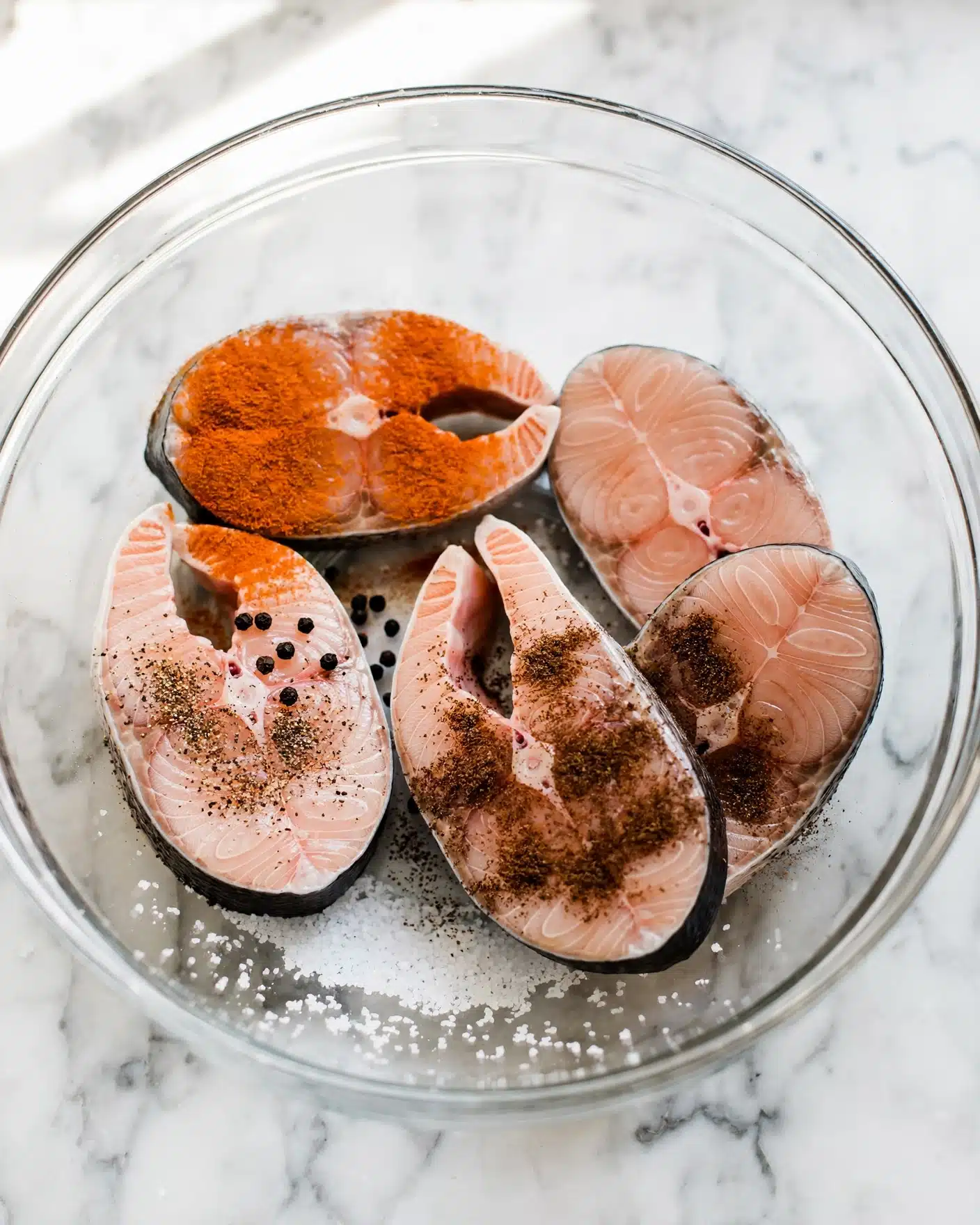 A clear glass bowl sits on a white marbled surface, holding six irregular chunks of raw fish with light pink flesh and some dark skin still attached. On the fish pieces, there are several piles of spices: bright yellow turmeric, ground black pepper, brownish powder, and fine white salt, all resting on top and around the fish, creating a contrast with the pale pink color. The bowl’s transparent sides show small water droplets inside, highlighting the freshness of the ingredients. photo taken with an iphone --ar 4:5 --v 7