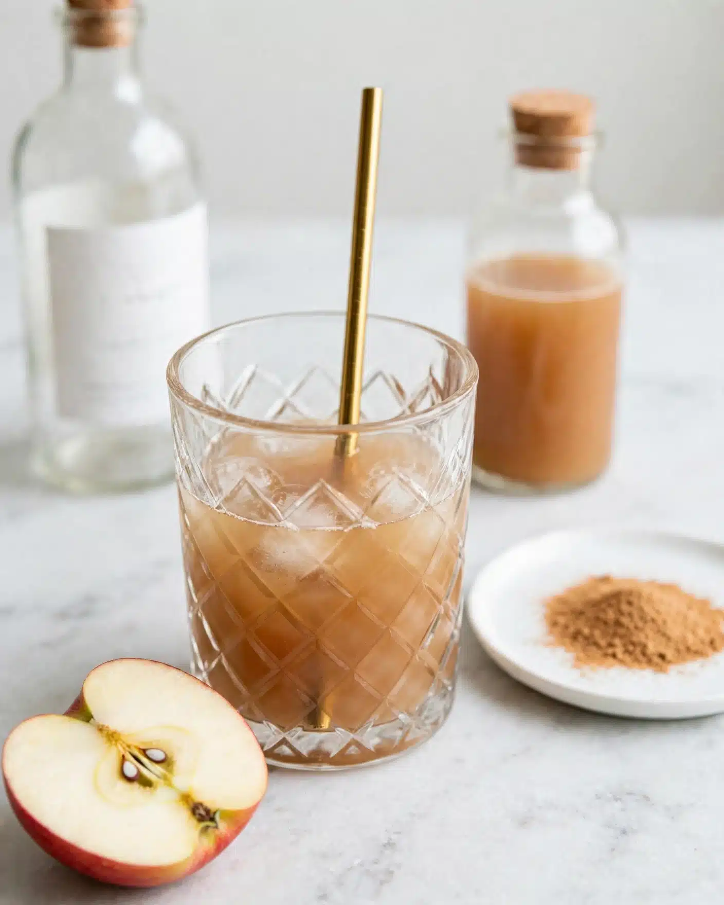 The Best Apple Cider Whiskey Cocktail Recipe 4 A clear glass with diamond patterns is filled about halfway with an orange-brown liquid, and a shiny gold metal straw is inside it, standing upright from the center. To the left of the glass, there is a clear bottle with a white label and black cap, filled partially with an amber liquid. In front of the drink on the white marbled surface, there is a thin slice of apple showing the seeds. On the right side of the glass, there is a small white plate with a light-colored powder and a clear bottle filled with a bright orange liquid. The background is a plain white marbled texture. Photo taken with an iphone --ar 4:5 --v 7