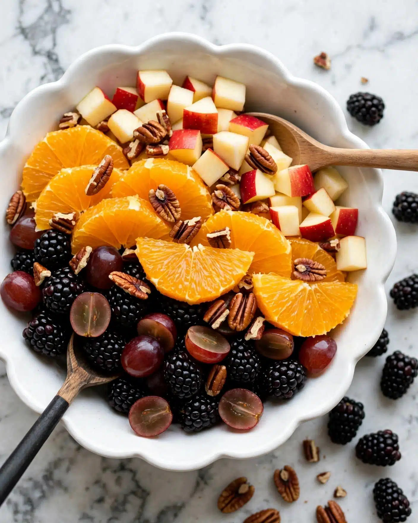 A white scalloped bowl holds a vibrant fruit salad with multiple layers: the bottom layer is dark blackberries with a bumpy texture, mixed with smooth, deep red grapes cut in halves. On top of this base, there are bright orange slices with a juicy texture, and small cubes of apple with red skin and creamy inside. Scattered among the fruits are crunchy brown pecan pieces. Two wooden spoons rest inside the bowl, one with a black handle visible in the lower left corner. The bowl sits on a white marbled surface, and some pecans and blackberries are scattered beside the bowl. photo taken with an iphone --ar 4:5 --v 7