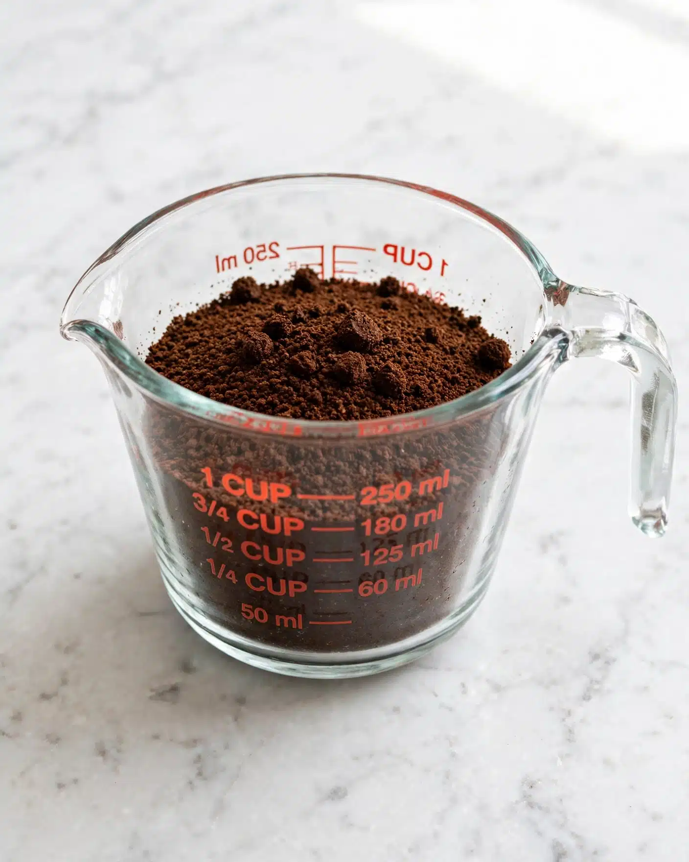 A clear glass measuring cup filled with a single layer of dark brown cocoa powder with small clumps scattered on top, placed on a white marbled surface. The measuring cup shows red measurement marks along the sides and has a spout and handle visible in the foreground. The cocoa powder fills the cup up to about one-third full, with a smooth but slightly uneven texture. Photo taken with an iphone --ar 4:5 --v 7