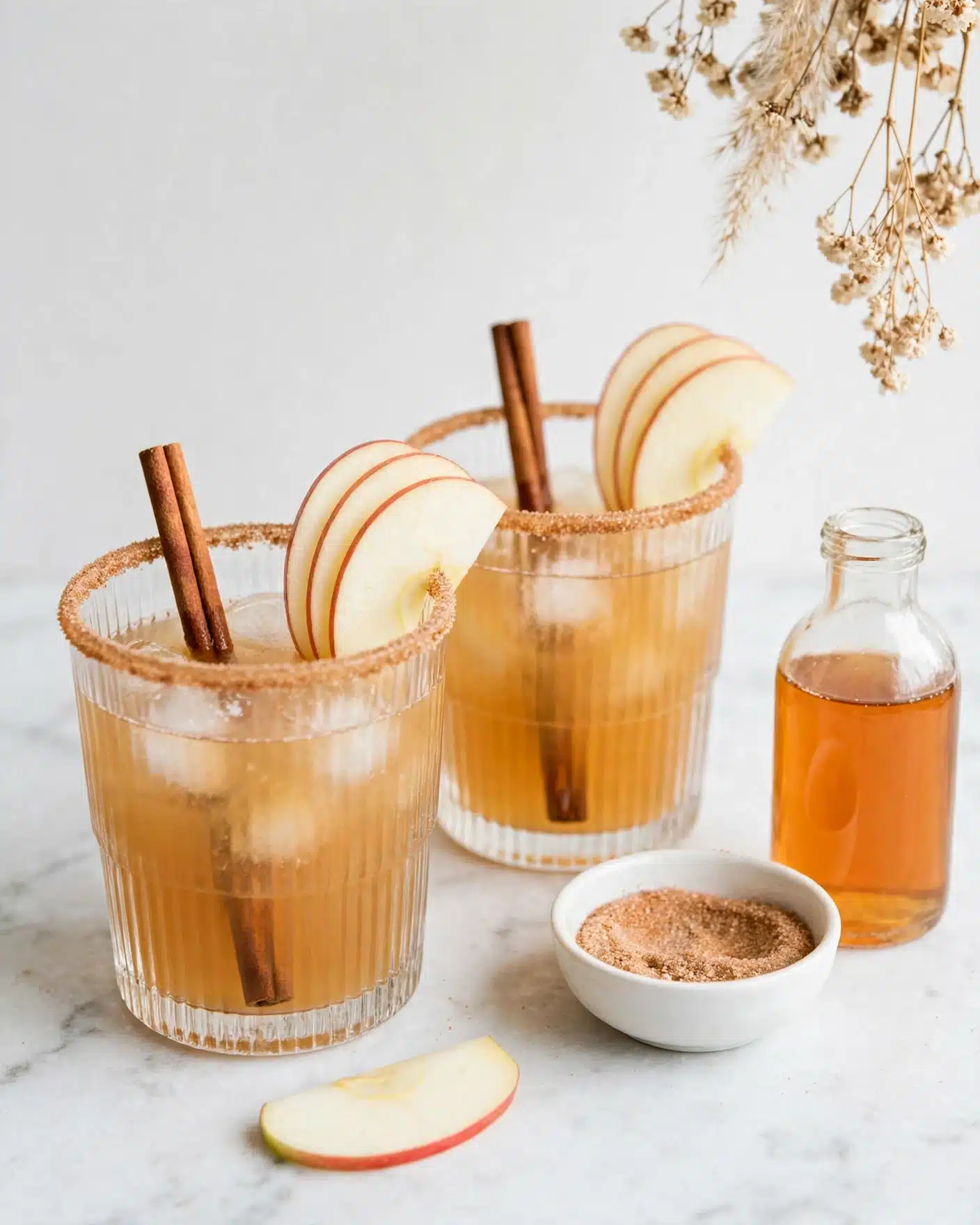 The Best Apple Cider Whiskey Cocktail Recipe 5 Two crystal-clear glasses are filled with a golden iced drink, each glass showing a textured diamond pattern. The top layer is pale and foamy with ice cubes visible inside the liquid. Each glass has three thin slices of pale yellow apple resting on the rim and a brown cinnamon stick standing upright inside the glass. The rims are coated with a rough, light brown sugar or spice mix. In the background, there is a clear glass bottle filled with amber liquid, and near the glasses a light beige bowl holds a fine powder, likely cinnamon sugar. The scene is set on a white marbled surface with a soft white backdrop and a dried flower spray to the right. Photo taken with an iphone --ar 4:5 --v 7