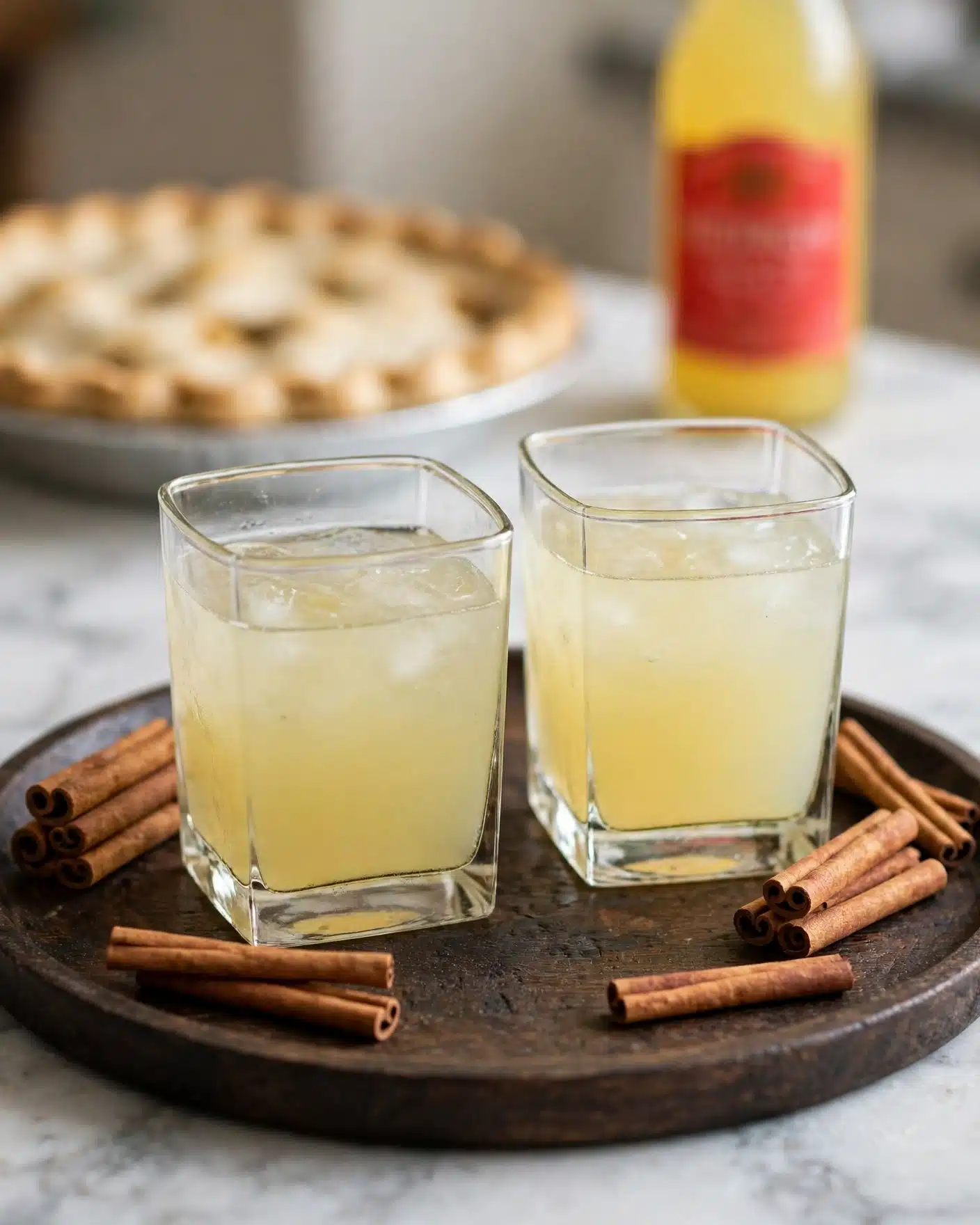 Two clear square glasses filled with a pale yellow drink sitting on a dark wooden tray. Around the glasses are several cinnamon sticks, some lying flat and some stacked. In the blurred background, there is a light-colored pie with a crimped edge and a yellow bottle with a red label beside it. The whole scene rests on a white marbled surface. photo taken with an iphone --ar 4:5 --v 7